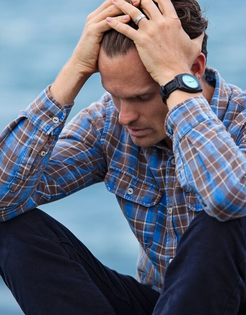 A man in a plaid shirt sits by the water looking distressed, symbolizing stress.