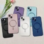 Six iPhone cases in black, pink, purple, white, teal, and blue with circular MagSafe rings, laid on a light surface.